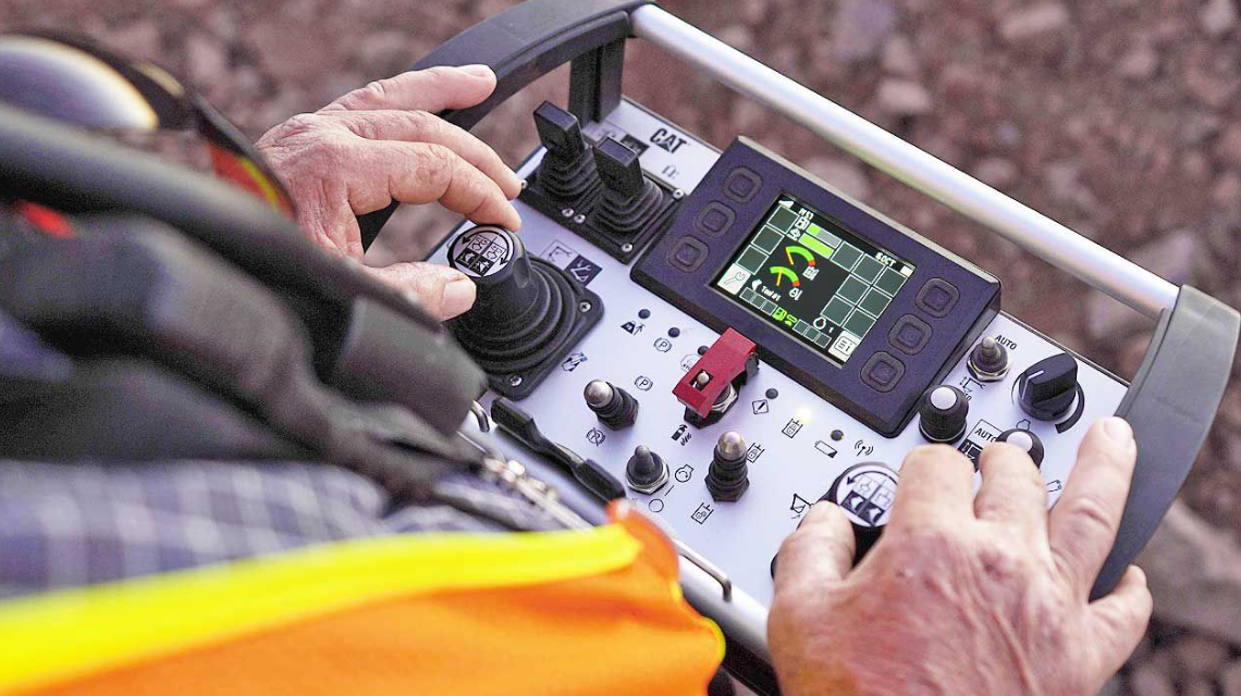 Close-up of an operator’s hands using a Cat Command remote control system with joystick and screen interface, showcasing advanced machine control technology.