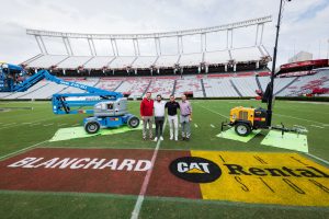 Jeremiah Donati (USC Athletic Director), standing next to Rozier Blanchard (Executive Vice President and CCO of Blanchard Machinery), Boyd Blanchard (President and CEO of Blanchard Machinery), and Shane Beamer (Head Football Coach of USC Gamecocks) on the football field at Williams Brice Stadium with Blanchard Rental Services logo painted on the field and Blanchard Rental equipment pictured in the background